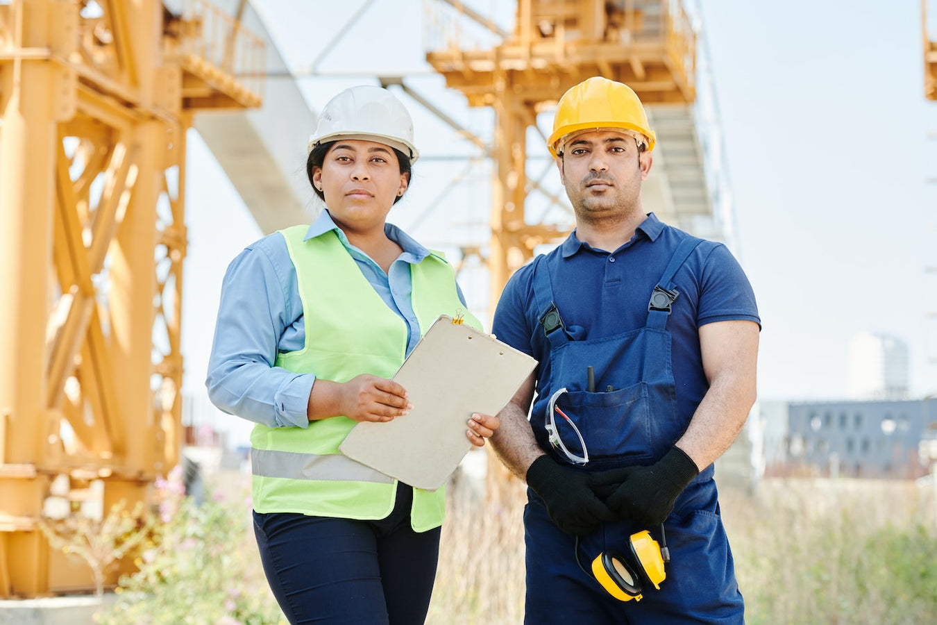 Two people standing at a construction site. On the left they are wearing a white hardhat, green safety vest, and holding a clipboard. On the right, they are wearing a yellow hardhat and a blue work apron.