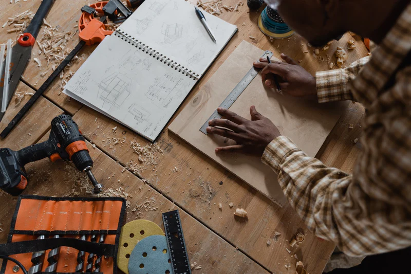 Top view of a work table showing power drill, notebook, and a man using a ruler.